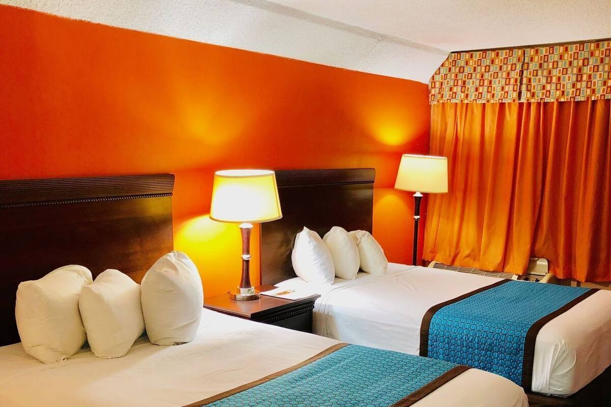 A hotel room with two beds, each with white bedding and a blue geometric-patterned throw. There are two bedside lamps, a wooden headboard, and a curtain with a geometric pattern.