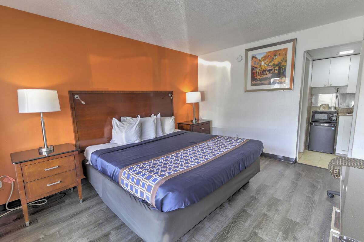 A hotel room with a large bed, two bedside tables with lamps, a small desk, a kitchenette, and a painting on the wall.