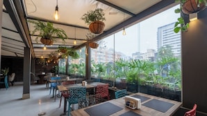 An indoor dining area with tables and chairs, hanging plants, and a view of buildings through large windows.