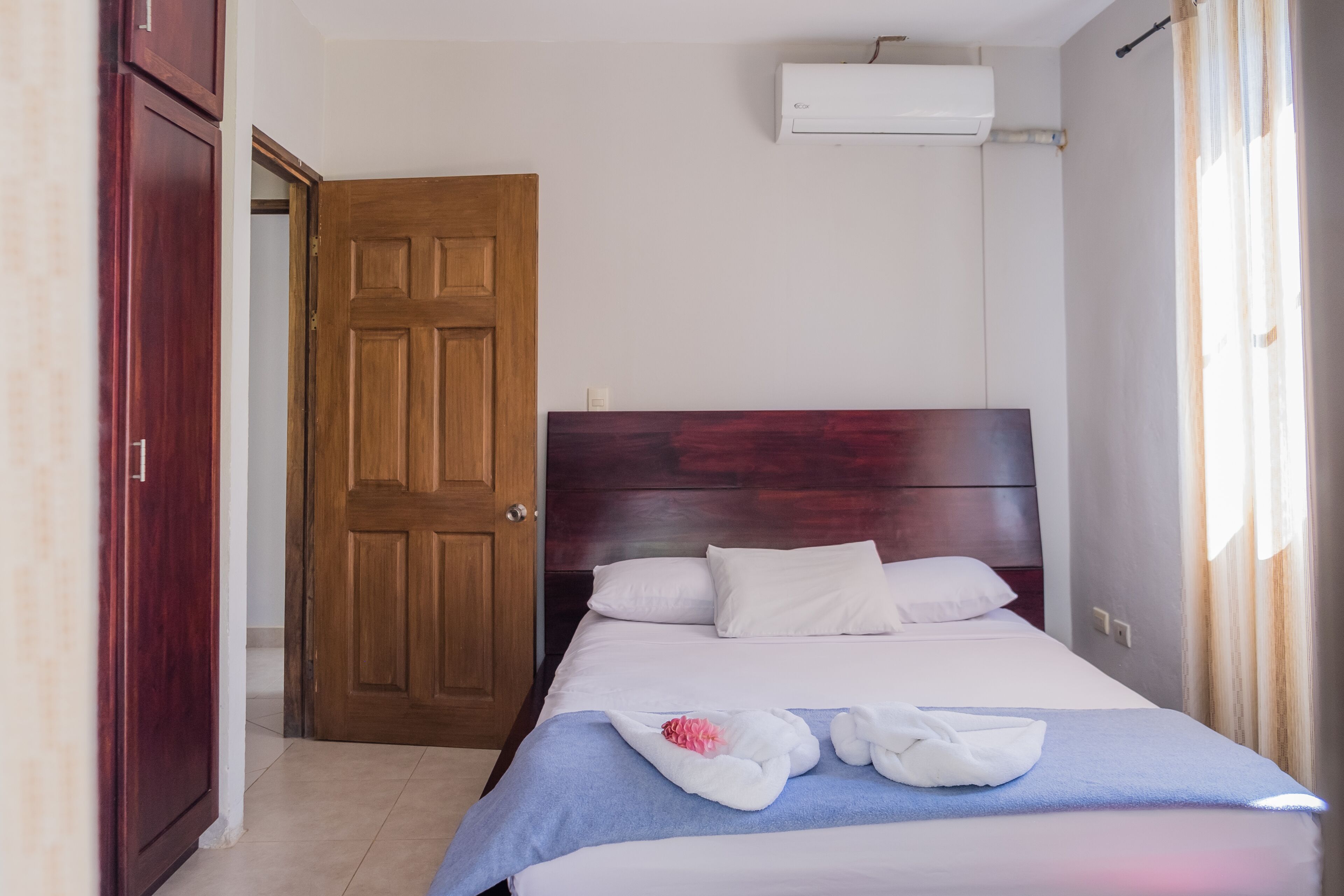 Ein Hotelzimmer mit Holztür, einem Bett mit weißer Bettwäsche und einer Klimaanlage an der Wand.