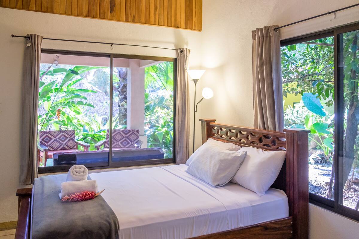 A bed with white linens, a wooden headboard, and a view of lush greenery through a window.