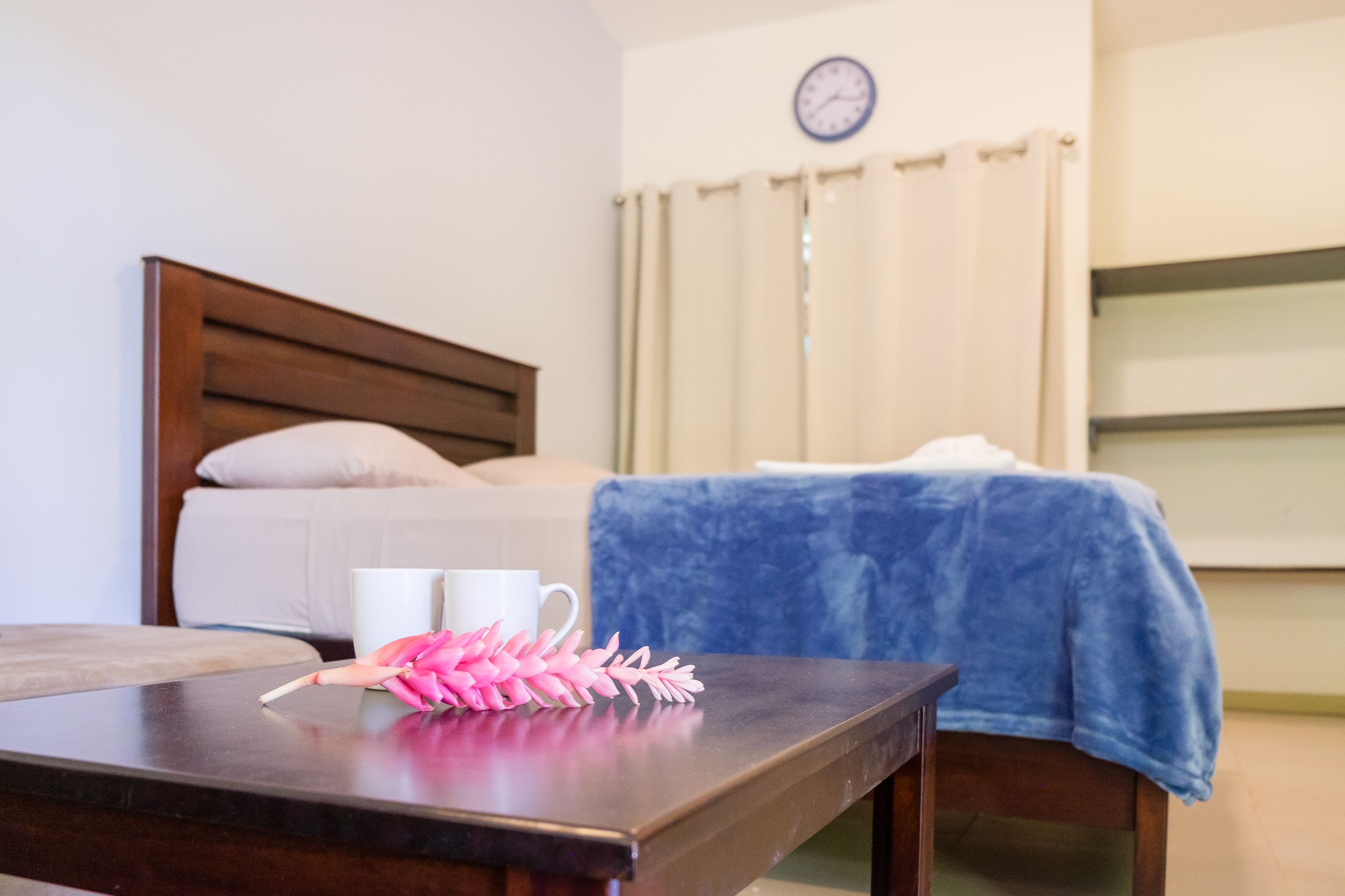 A bedroom with a bed, a bedside table with a cup and a decorative item, a clock on the wall, and a window with curtains.