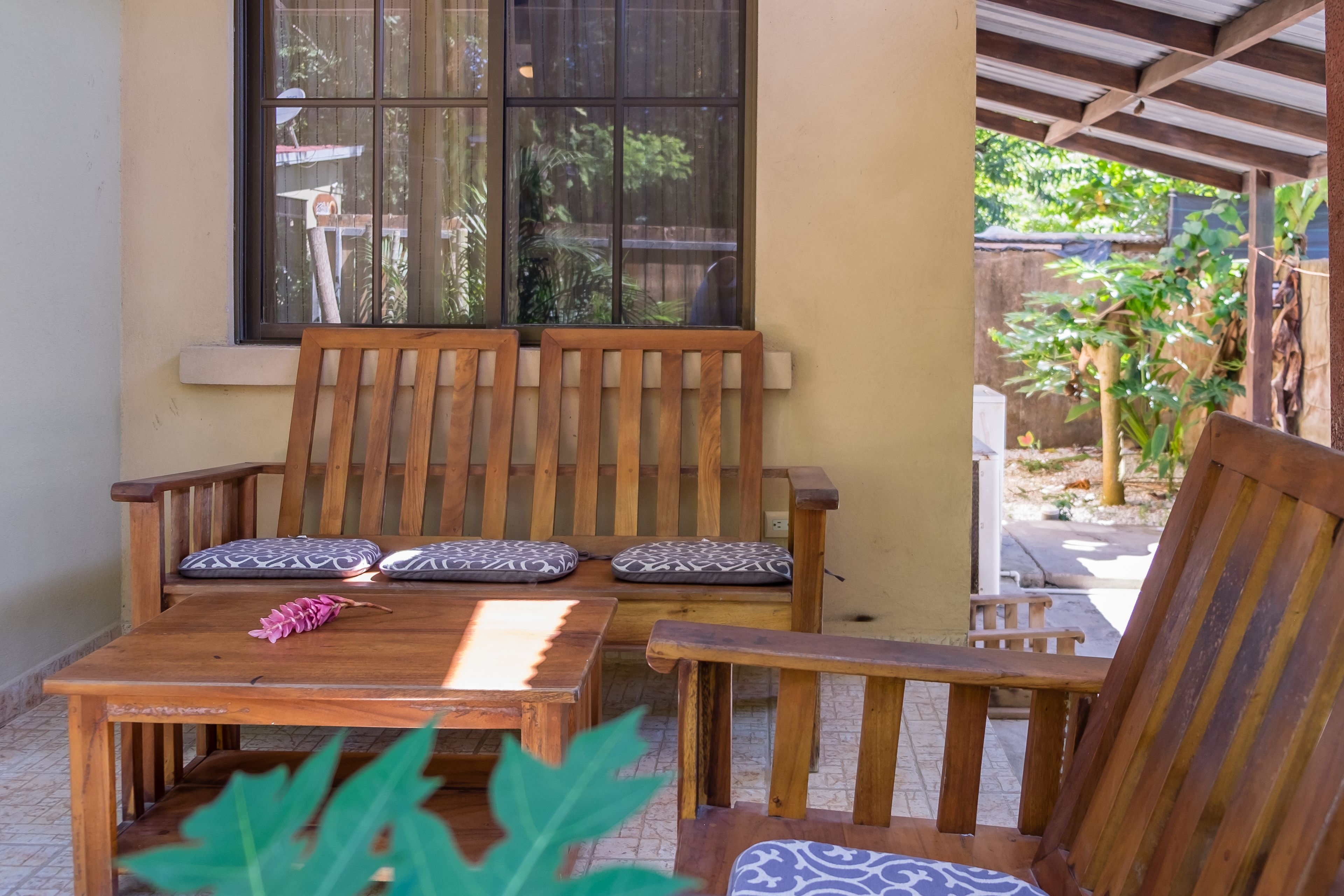 Un banco de madera con cojines estampados, una mesa de madera y una ventana con vista a la vegetación.