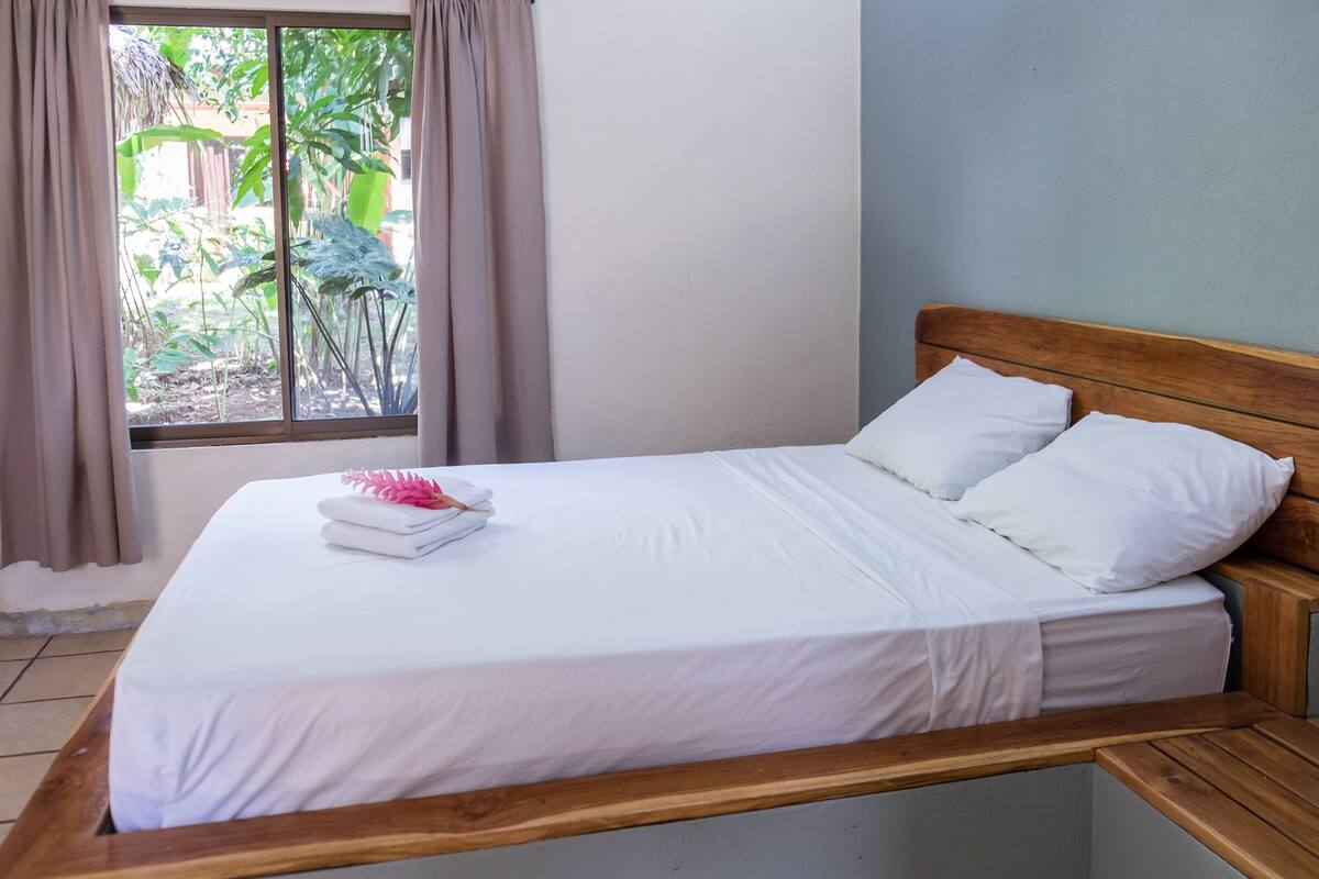 A neatly made bed with white linens and pillows, a wooden headboard, and a window with curtains.