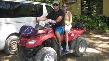 Een man en een vrouw op een rode terreinquad met een paarse rugzak, geparkeerd naast een witte bus.