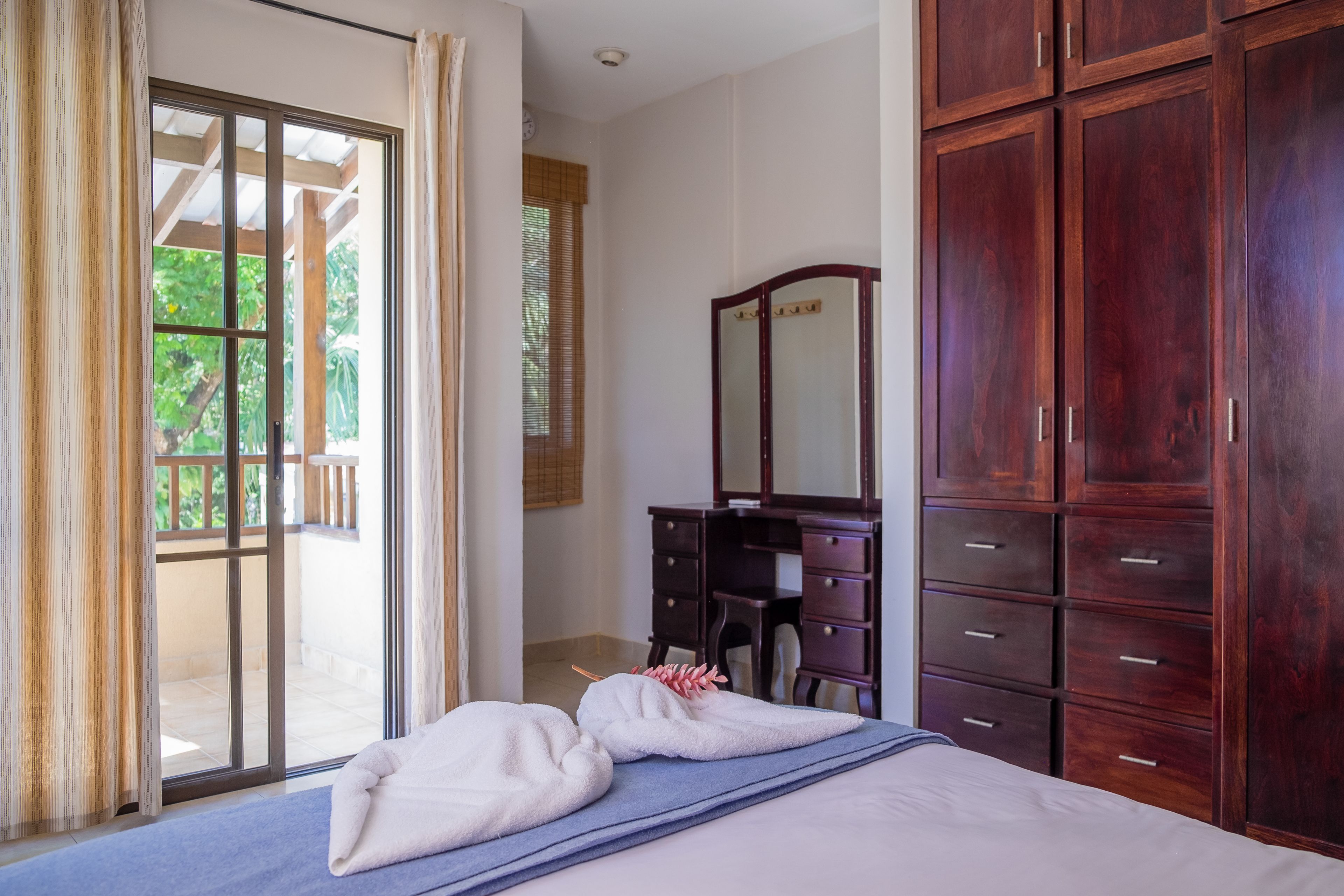 A bedroom with a bed, a wooden wardrobe, a dresser, and a sliding glass door leading to an outdoor area.