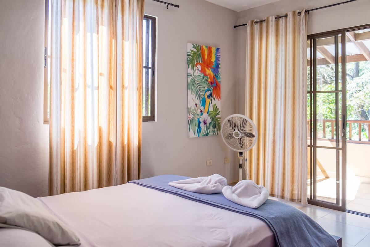 A hotel room with a bed, a fan, and a painting of tropical birds.