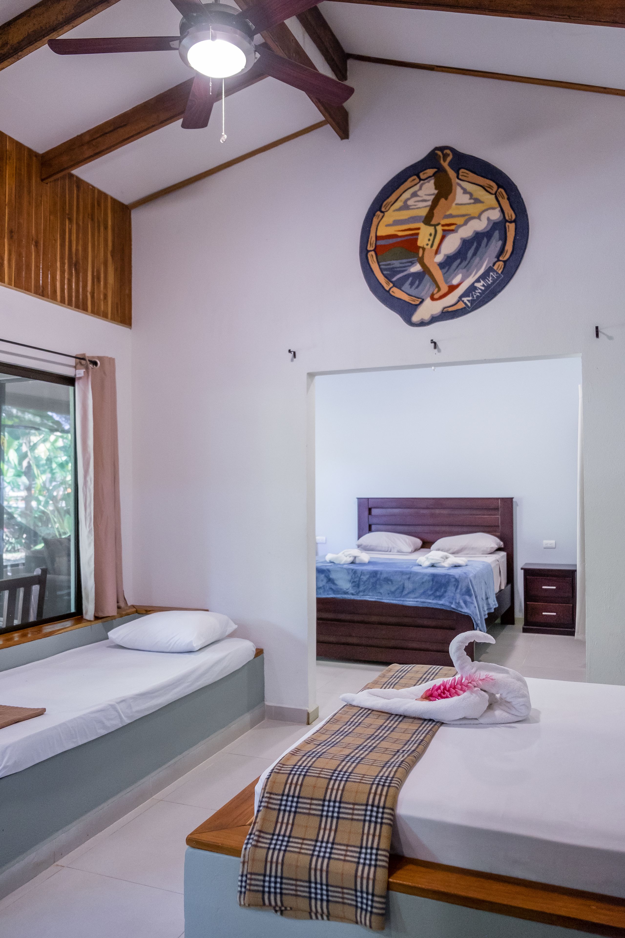 Un dormitorio con dos camas individuales, un ventilador de techo de madera, un espejo grande y un adorno decorativo en la pared.