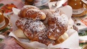 Een mand met gebak, waaronder croissants, en een koffiekop op een tafel met een bloemrijk tafelkleed.