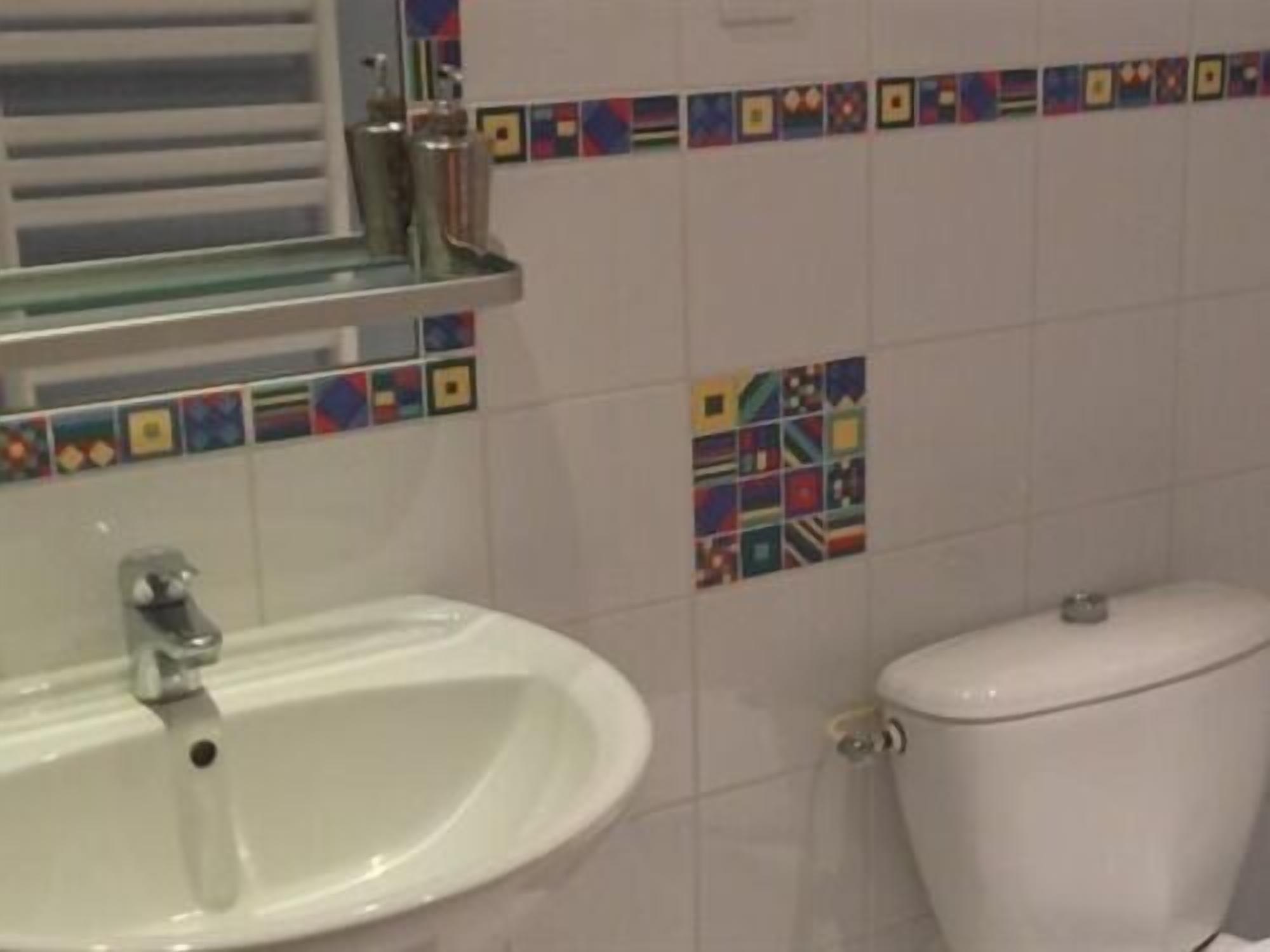 A bathroom with a white sink, a toilet, and a mirror above the sink. The walls are tiled with a colorful border design.