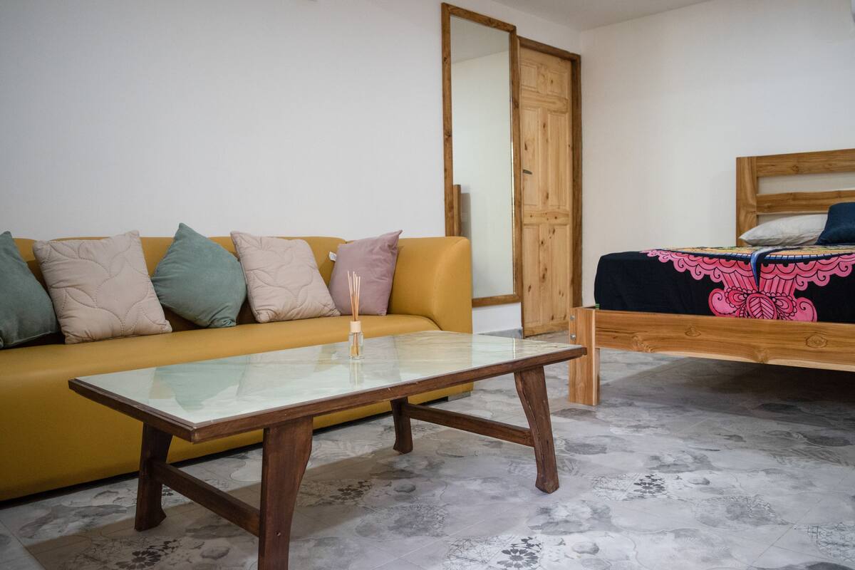 Een kamer met een gele bank, een houten bed met een zwartroze dekbedovertrek en een salontafel met een glazen blad, en een grote spiegel aan de muur.