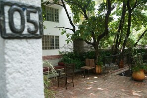 A patio area with a brick floor, a white wall with a number '05' sign, and a tree providing shade. - Corail Suites (Petionville)