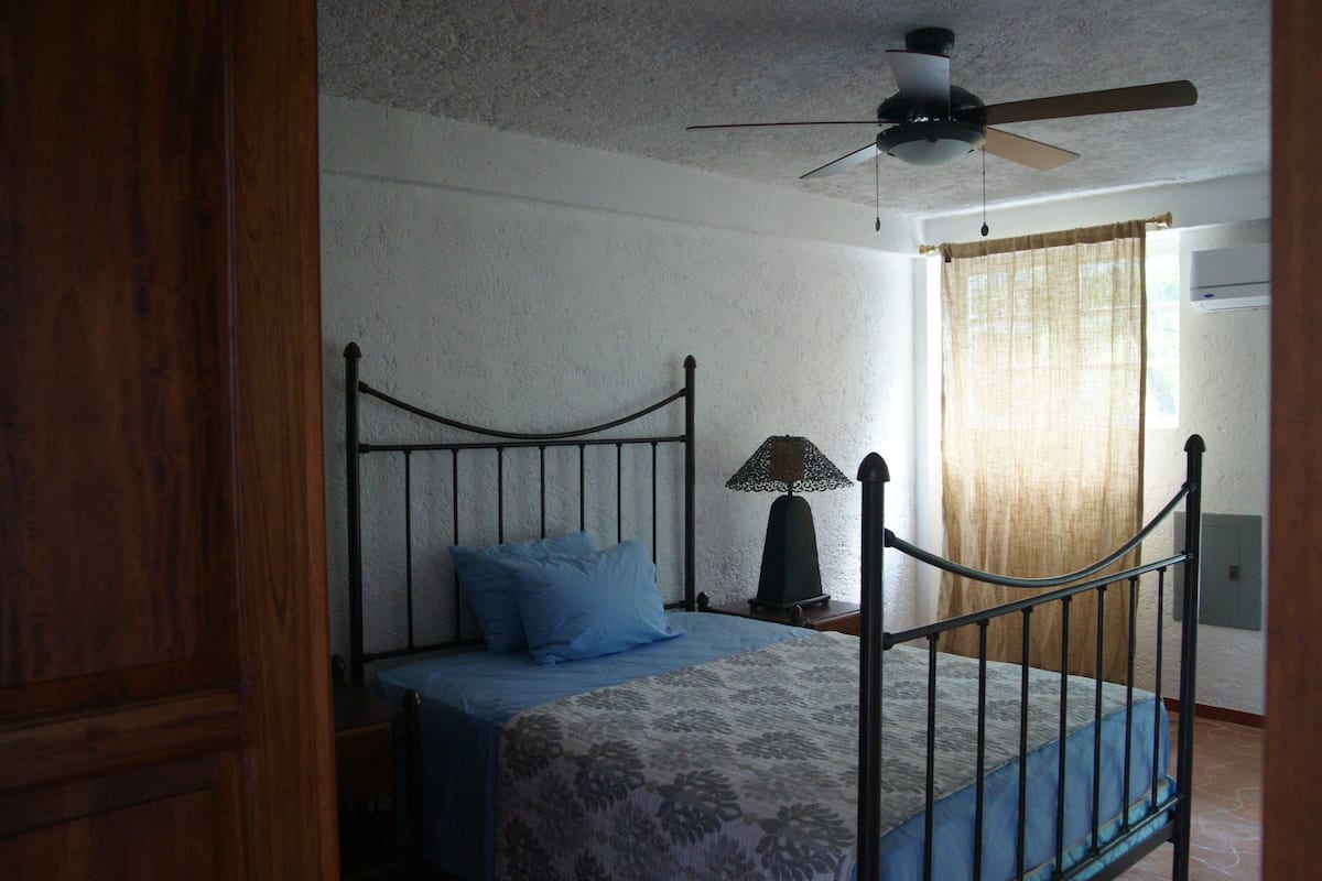Un dormitorio con una cama individual, ventilador de techo, mesita de noche y ventana con cortinas.