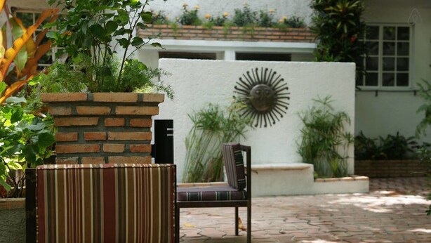 A patio area with a striped sofa and a chair, a brick planter with greenery, and a decorative wall with a sun motif. - Corail Suites (Petionville)