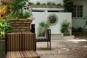 Un patio con un sofá a rayas y una silla, una jardinera de ladrillo con follaje y una pared decorativa con un motivo solar.