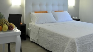 Quarto de hotel com uma cama grande, roupa de cama branca, uma mesinha com frutas e duas luminárias na parede.