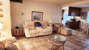 A living room with a beige sofa, a glass coffee table, and a wicker ottoman. There is a framed picture on the wall and a lamp on a side table. - Silver Sands Motel (Clearwater Beach)