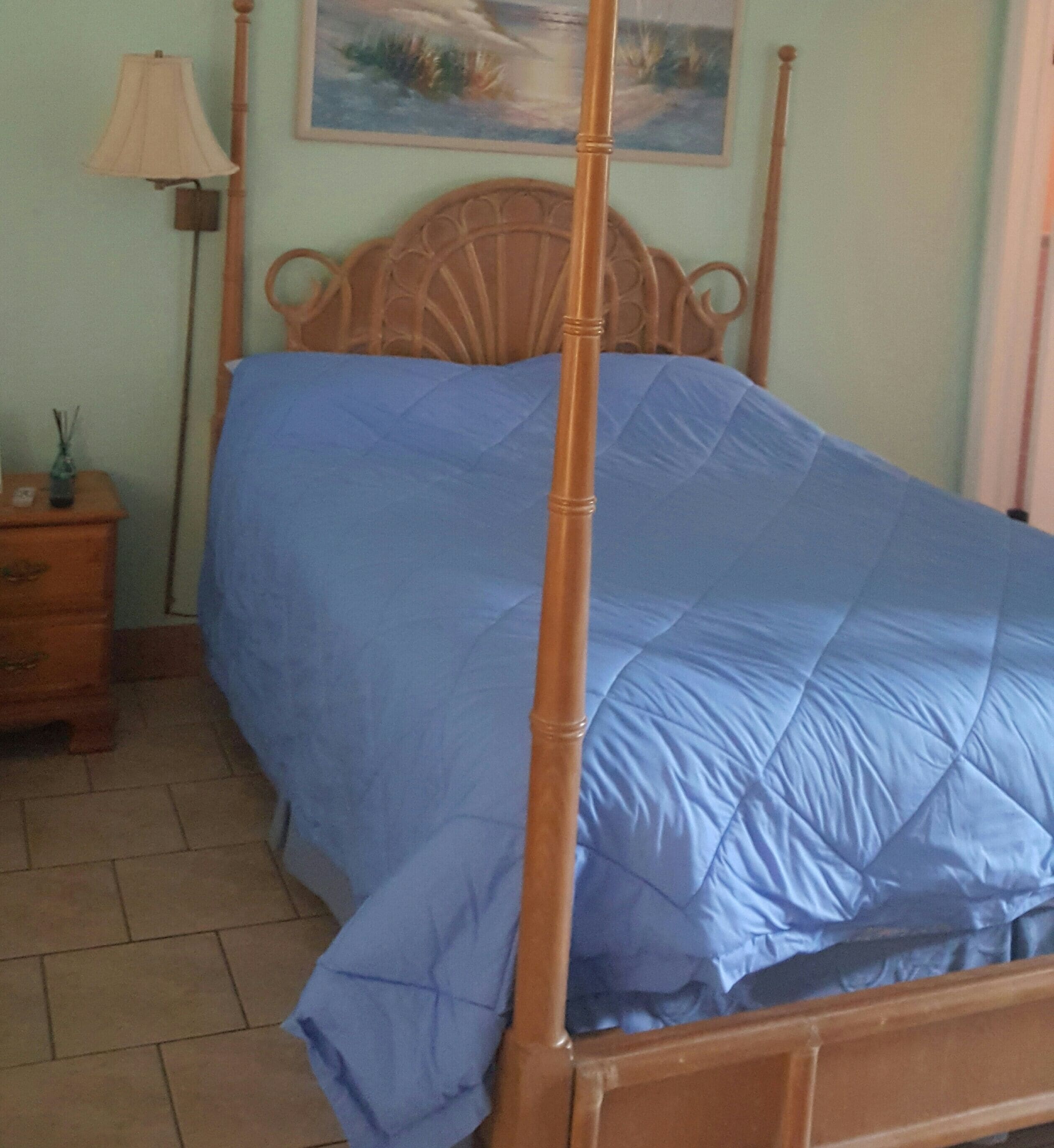Een bed met een blauw sprei, een houten hoofdbord, een nachtkastje met een lamp en een schilderij aan de muur.