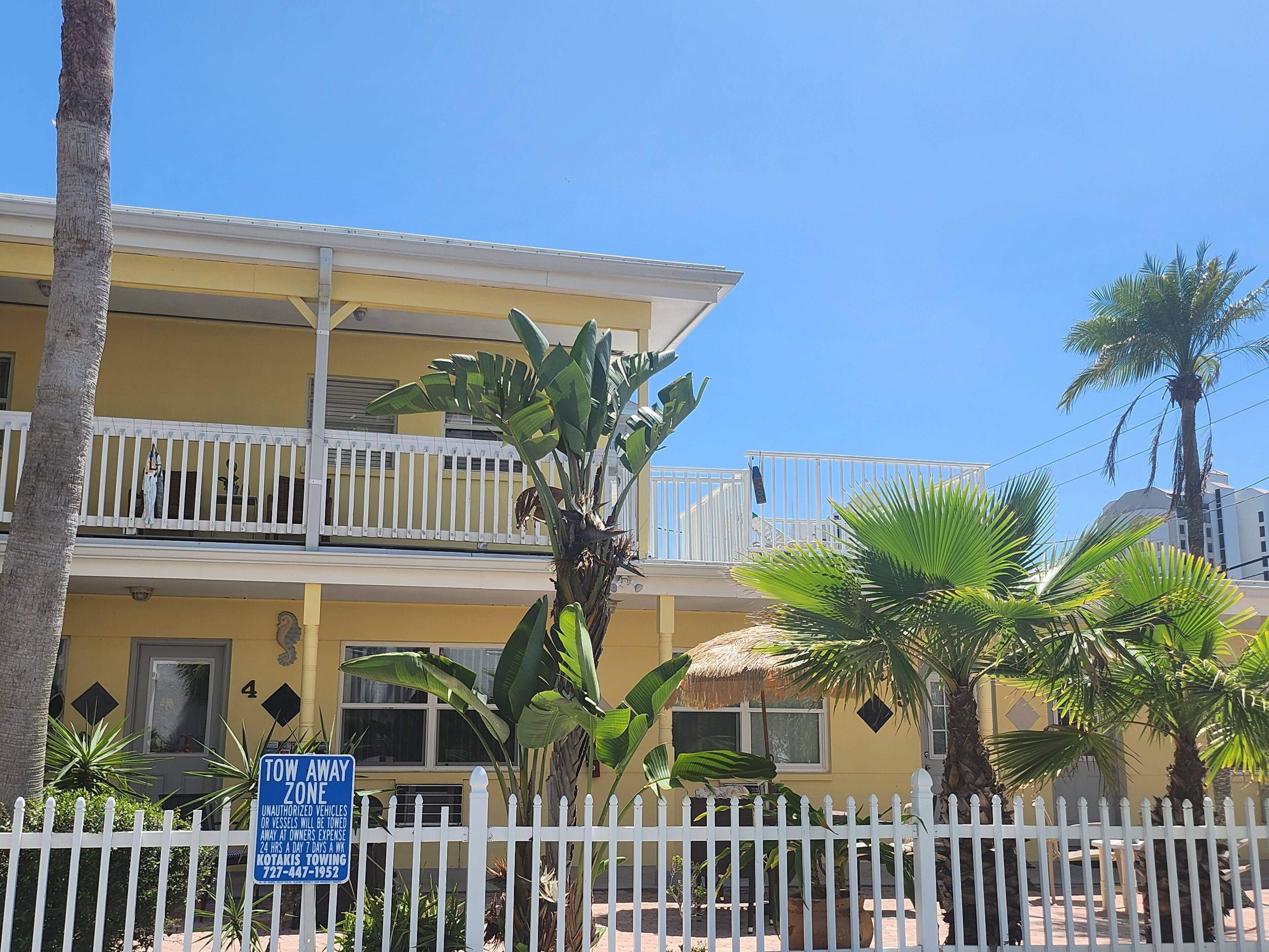 A yellow building with balconies, a white fence, and tropical plants.