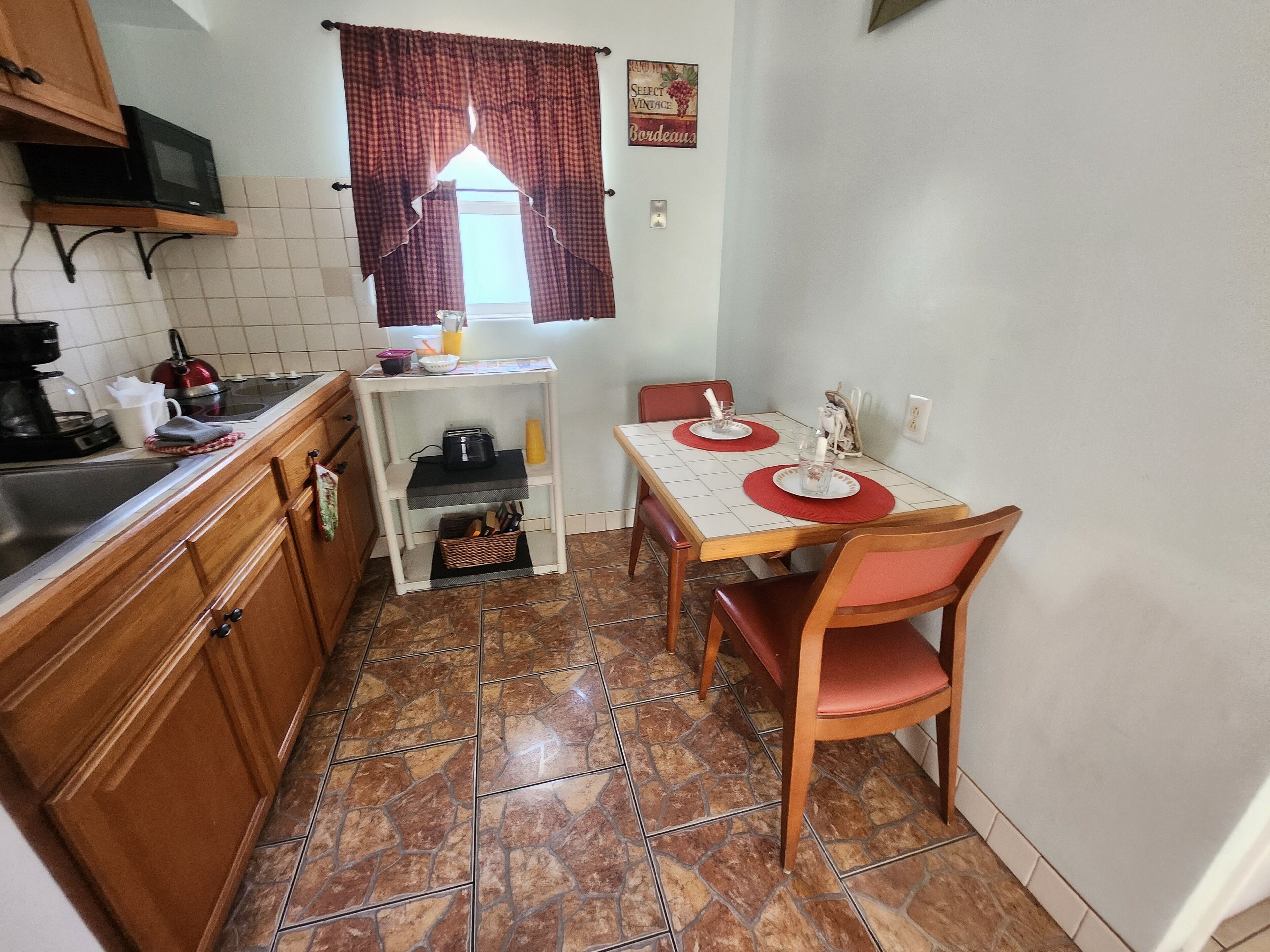 Une cuisine compacte avec un coin repas, dotée d’un sol carrelé, de meubles en bois, d’un four à micro-ondes, d’une bouilloire et d’une petite table avec des chaises.