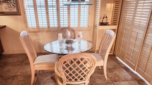 A dining area with a round glass table surrounded by wicker chairs, a window with blinds, and a tiled floor. - Silver Sands Motel (Clearwater Beach)