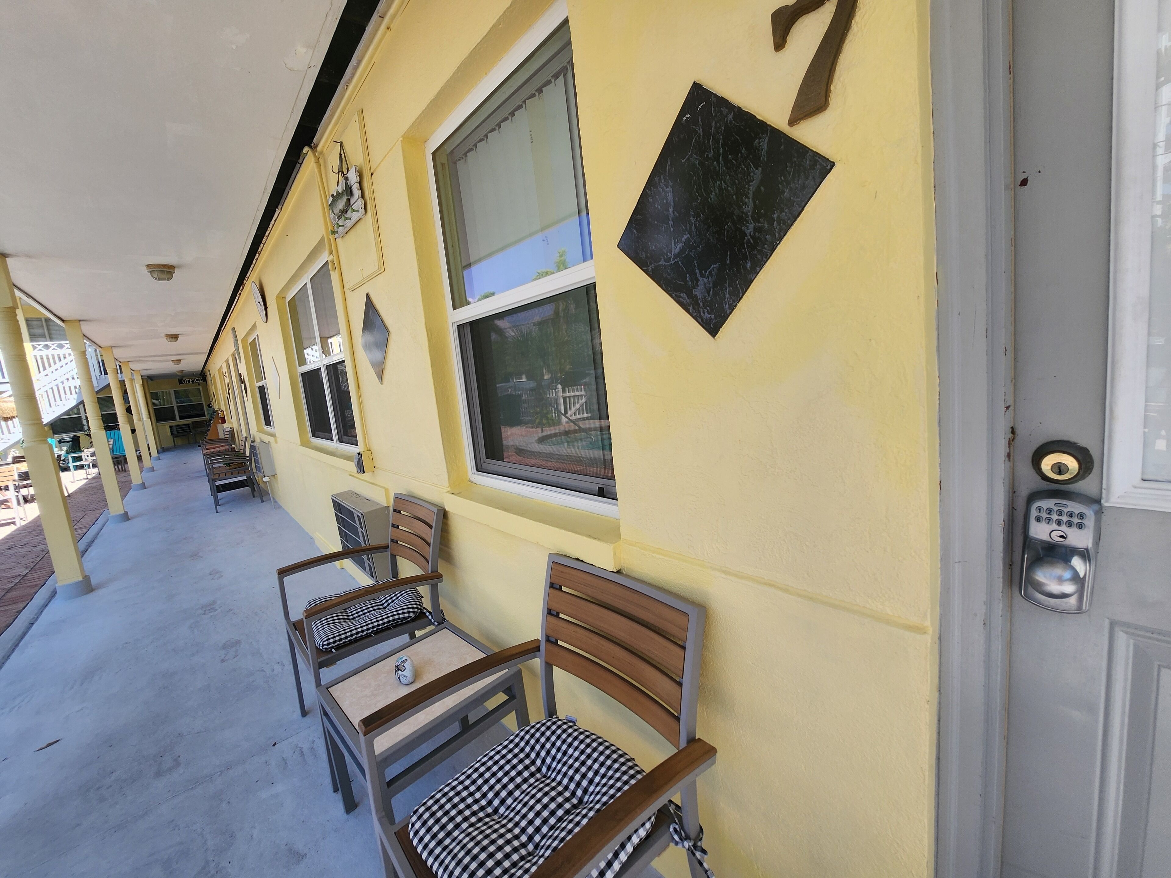 A yellow building with a covered walkway, outdoor seating with metal chairs and checkered cushions, and a door with a keypad lock.