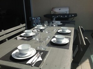Une table à manger dressée pour six personnes, avec des bols, des assiettes et des couverts blancs, et un barbecue en arrière-plan.