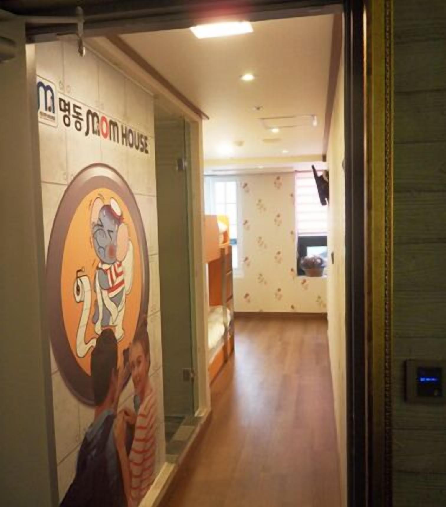 Un couloir avec une pancarte indiquant « M. House » et un personnage de dessin animé. On aperçoit un lit et une fenêtre avec des stores au fond.