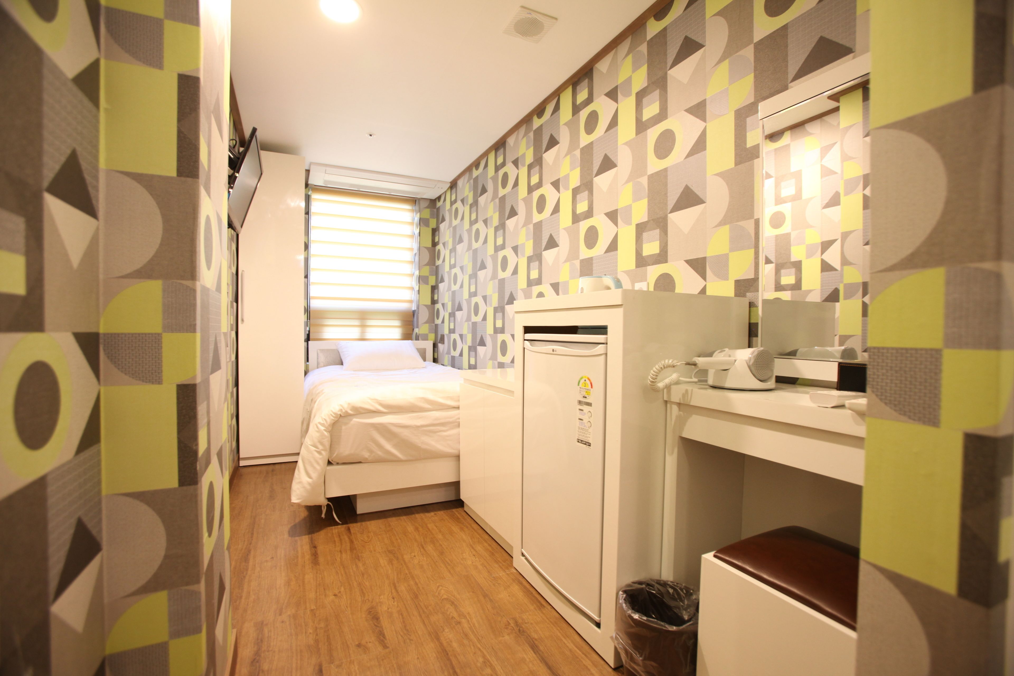 A compact hotel room with a bed, a small kitchenette, a refrigerator, and a trash bin.