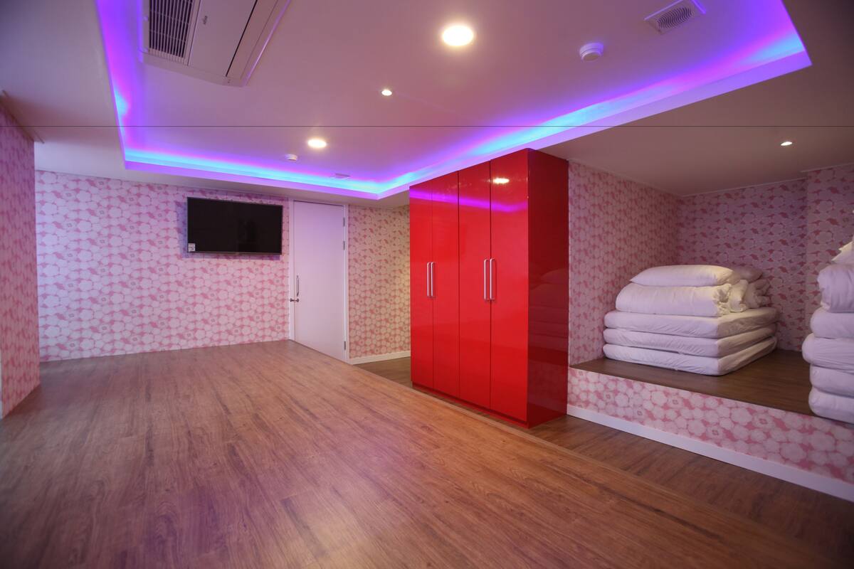Una habitación con suelo de madera, un armario rojo, un televisor de pantalla plana montado en la pared y una pila de toallas blancas.