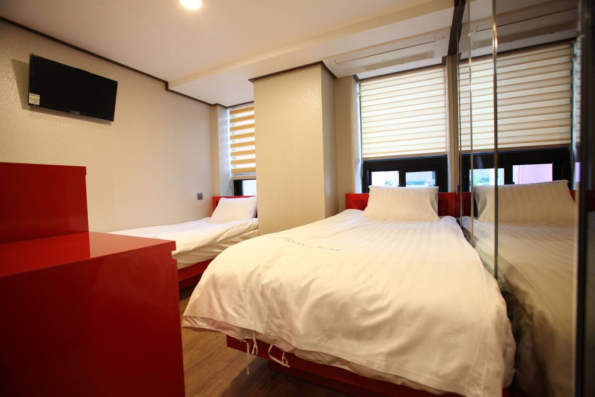 Habitación de hotel con dos camas individuales, un escritorio rojo, televisor montado en la pared y ventanas con persianas.