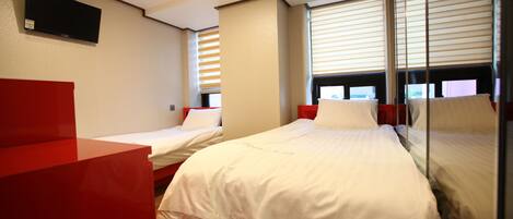Habitación de hotel con dos camas individuales, un escritorio rojo, televisor montado en la pared y ventanas con persianas.