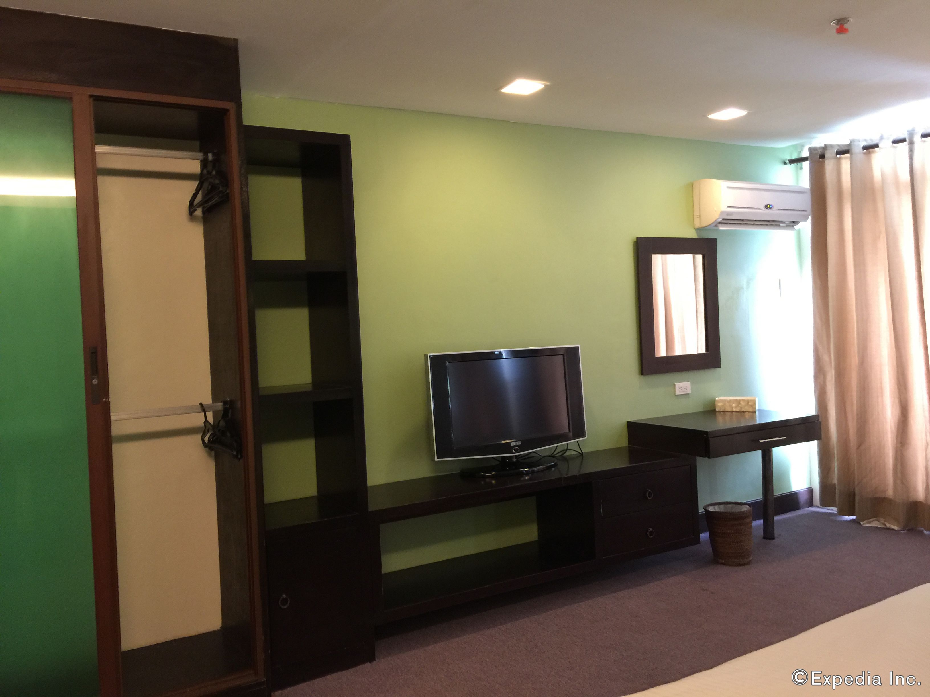 Una habitación con una televisión sobre un soporte de madera oscura, una mesita con una lámpara, un espejo y un ventanal con cortinas.
