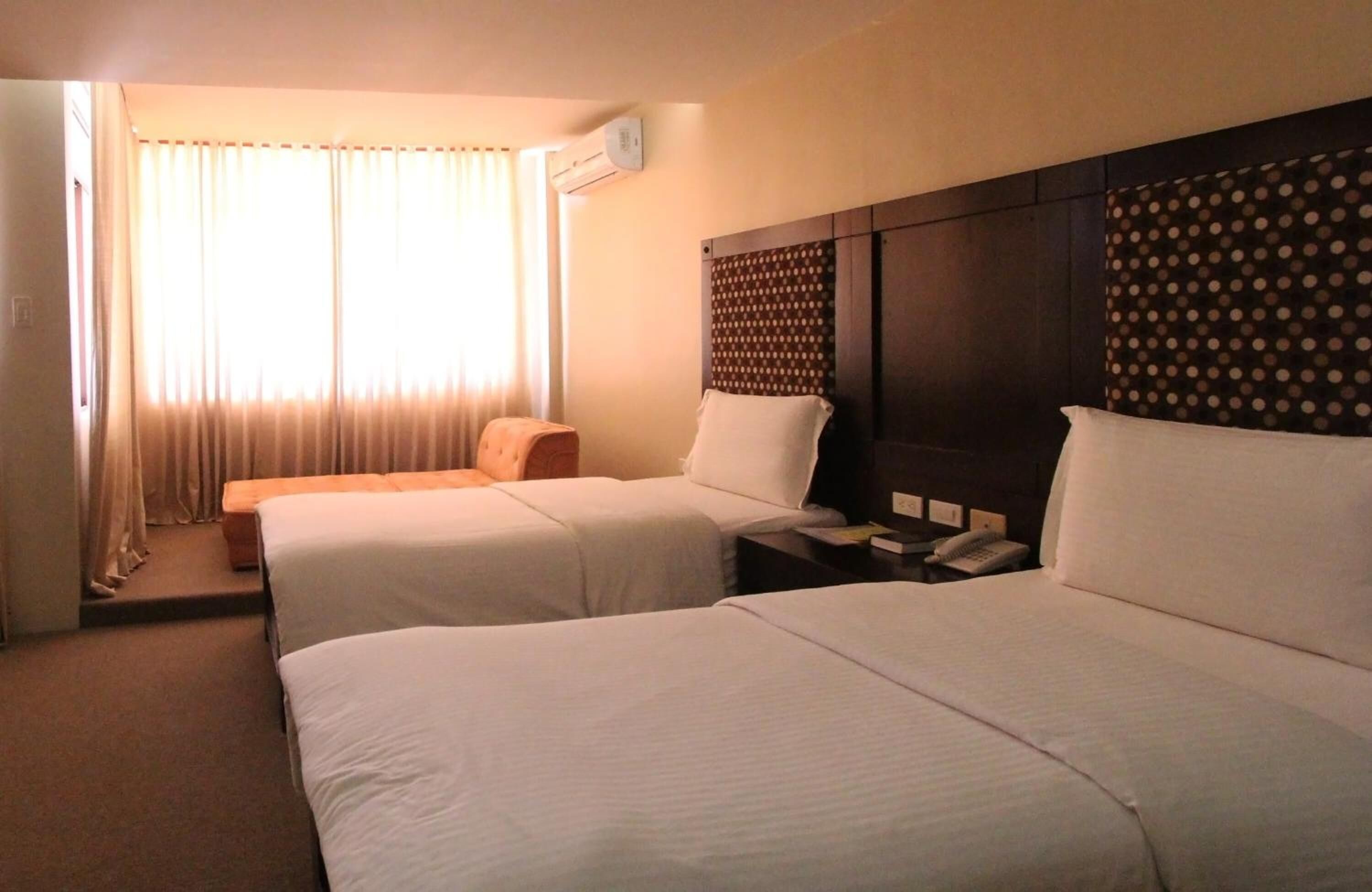 A hotel room with two single beds, a small sofa, a nightstand with a phone, and a window with curtains.
