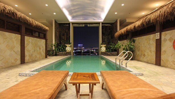 A pool area with lounge chairs and a table, a view of the city at night, and a thatched roof design. - Harmoni Suites Hotel (Batam)