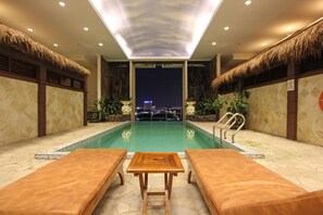 Zona de piscina con sillones de descanso y una mesa, vista nocturna de la ciudad y techo de paja.