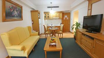 Una sala de estar con un sofá amarillo, una mesa de centro con fruta, un televisor y una zona de cocina con armarios de madera.