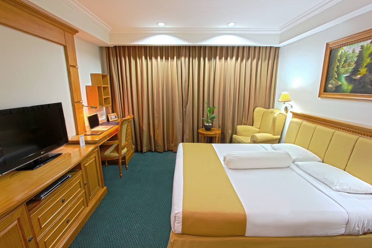 Una habitación de hotel con cama, escritorio con televisor, silla, lámpara y un cuadro en la pared.