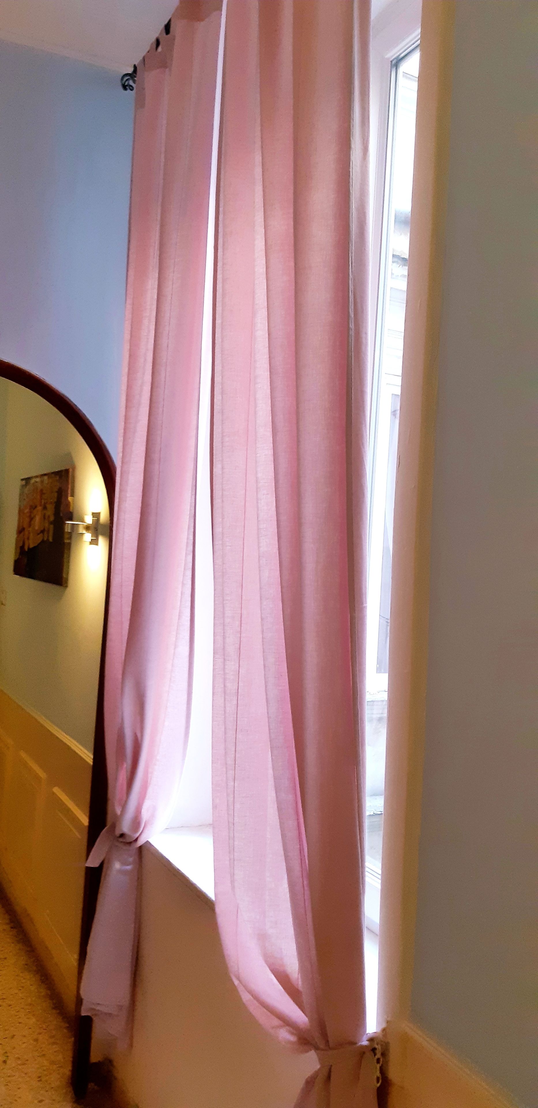 Un pasillo con una ventana cubierta por cortinas rosas, un espejo y una lámpara fijada a la pared.