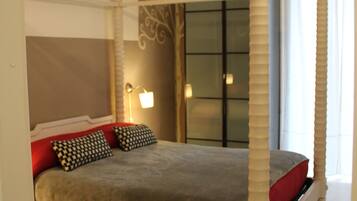 Una cama con dosel y cuatro columnas, con una colcha rojo y gris, dos almohadas estampadas y una mesita de noche con lámpara.