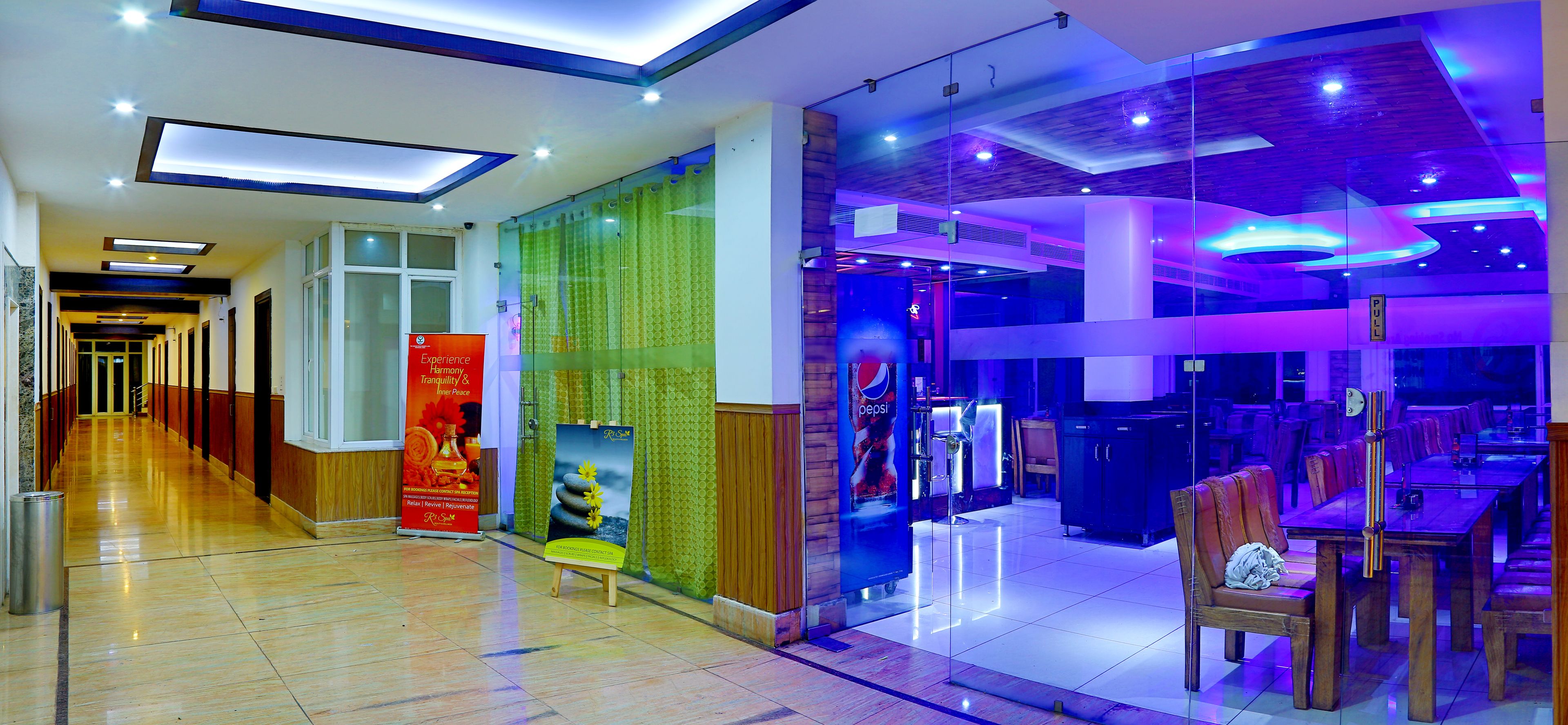 A hotel lobby with a long corridor, a glass partition with a Pepsi advertisement, and a dining area with tables and chairs.