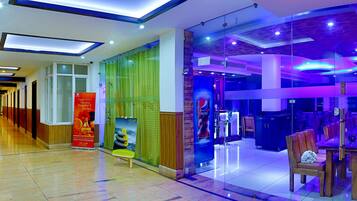 A hotel lobby with a long corridor, a glass partition with a Pepsi advertisement, and a dining area with tables and chairs.
