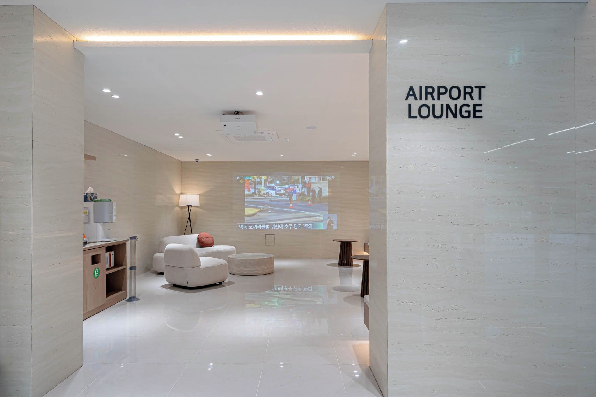 An airport lounge with a modern design, featuring a sofa, a lamp, and a projection screen displaying a news broadcast.