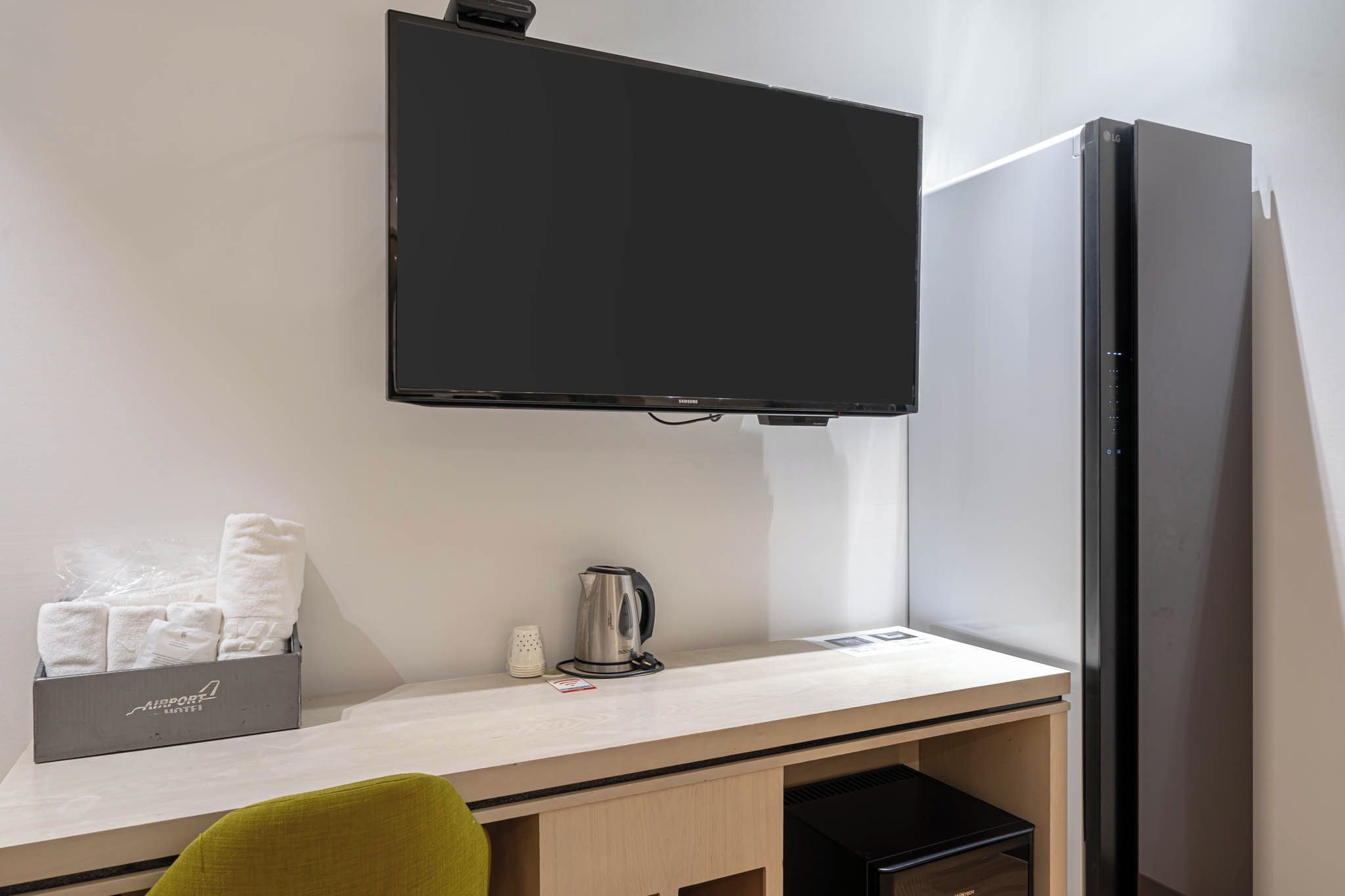 A hotel room with a flat-screen TV mounted on the wall, a small desk with a kettle, and a cabinet with towels.