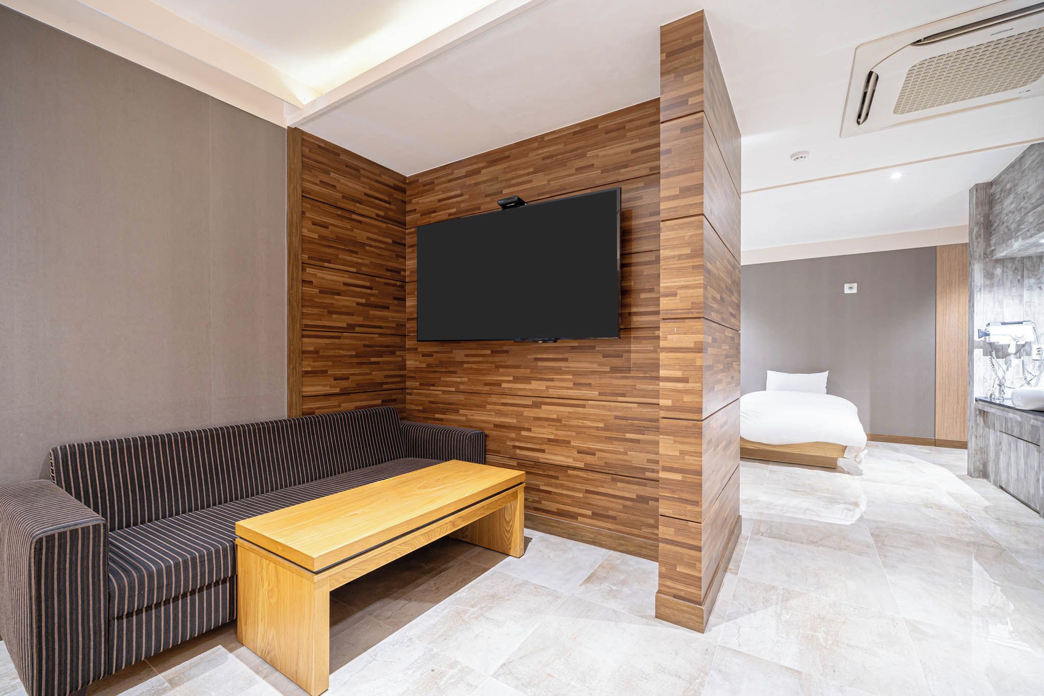 A modern hotel room with a sofa, a wooden coffee table, a flat-screen TV mounted on a wooden wall, a bed with a headboard, and a bathroom with a mirror and hairdryer.