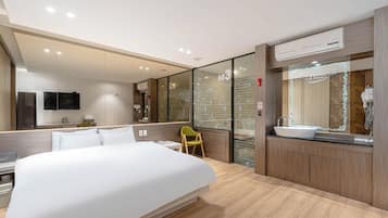 A modern hotel room with a large bed, a seating area, a bathroom with a sink, and a glass wall with a number.