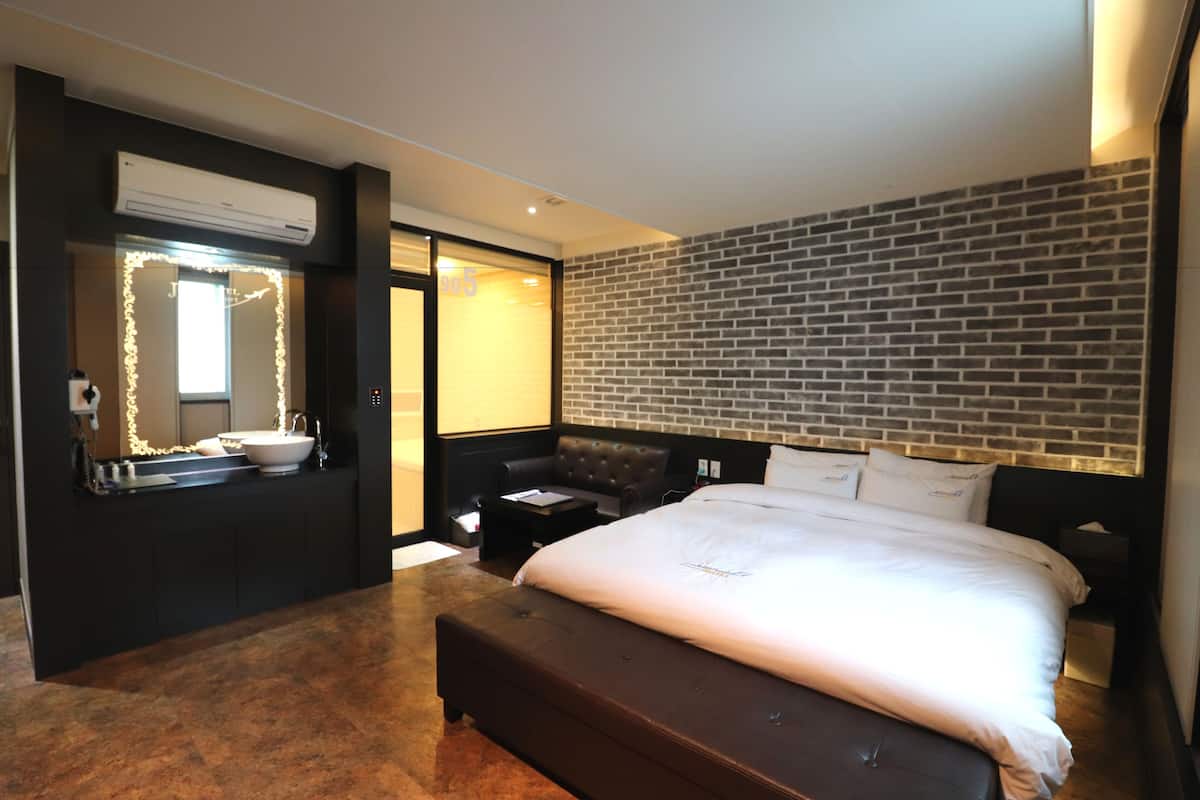 A modern hotel room with a large bed, a built-in vanity with a mirror, a brick-patterned headboard, and a small seating area with a leather couch.