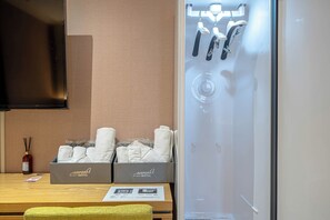 A hotel room with a wooden table, two boxes of towels, a bottle of air freshener, and a glass shower enclosure. - Airport Hotel (Busan)