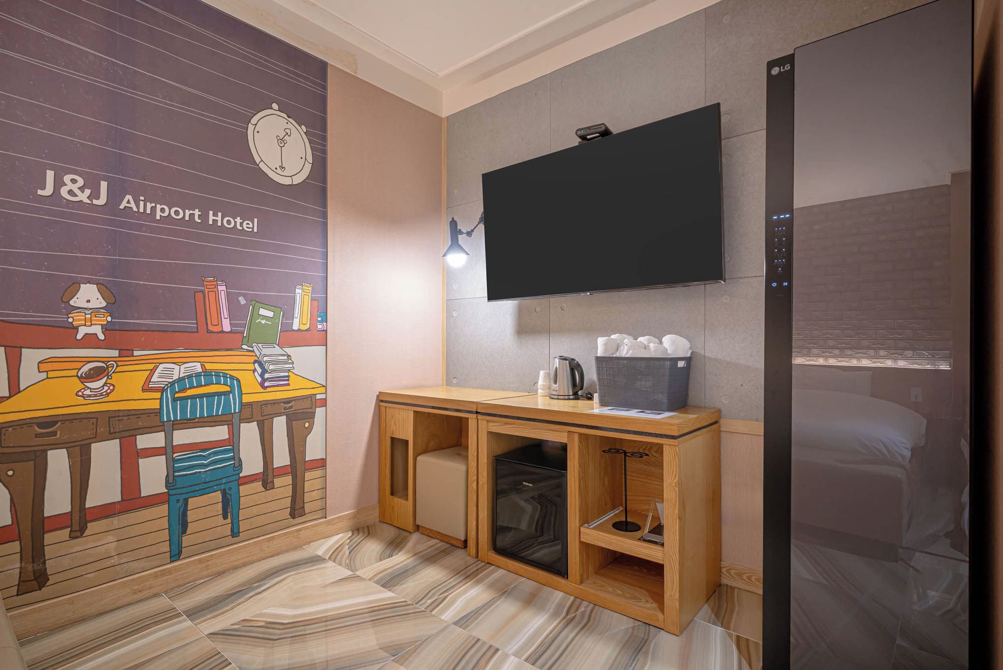A hotel room with a large flat-screen TV mounted on the wall, a wooden console table with a kettle and a basket, and a mural depicting a cozy reading nook.