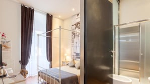 A bedroom with a four-poster bed, a bedside table with a lamp, a wardrobe, and a bathroom with a shower and toilet. - Vacanze Romane Rooms (Rome)